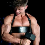 Muscular man lifting a dumbbell with focused expression, highlighting bicep, chest, and tricep muscles against a dark gym background.